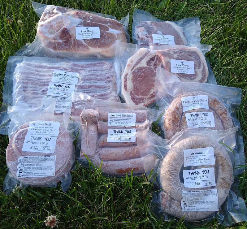 Assorted vacuum-sealed pork products from Country View Farm, including pork chops, bacon, smoked kielbasa, bratwurst, and pork roast, displayed on grass.