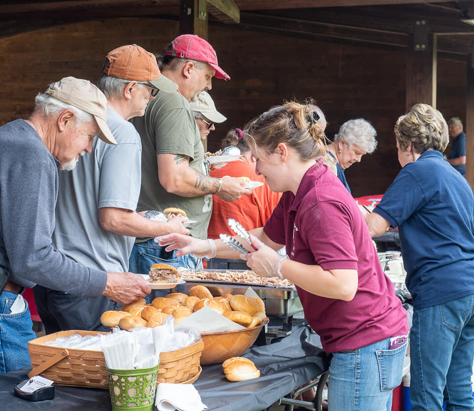 Group of people enjoying a community meal at a picnic, with a woman serving food from a buffet table filled with rolls and pulled pork. The setting features a rustic wooden pavilion, highlighting the social atmosphere of the event.