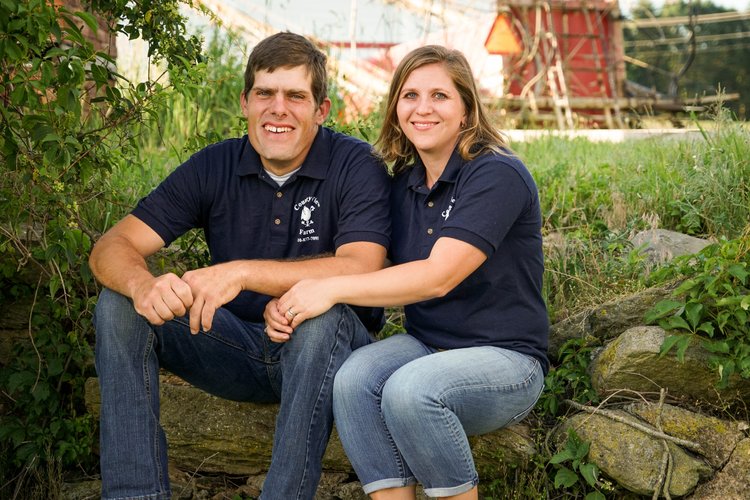 Smiling couple sitting on a rock in a green field, wearing matching navy blue polo shirts, with a rustic farm backdrop featuring a red barn.