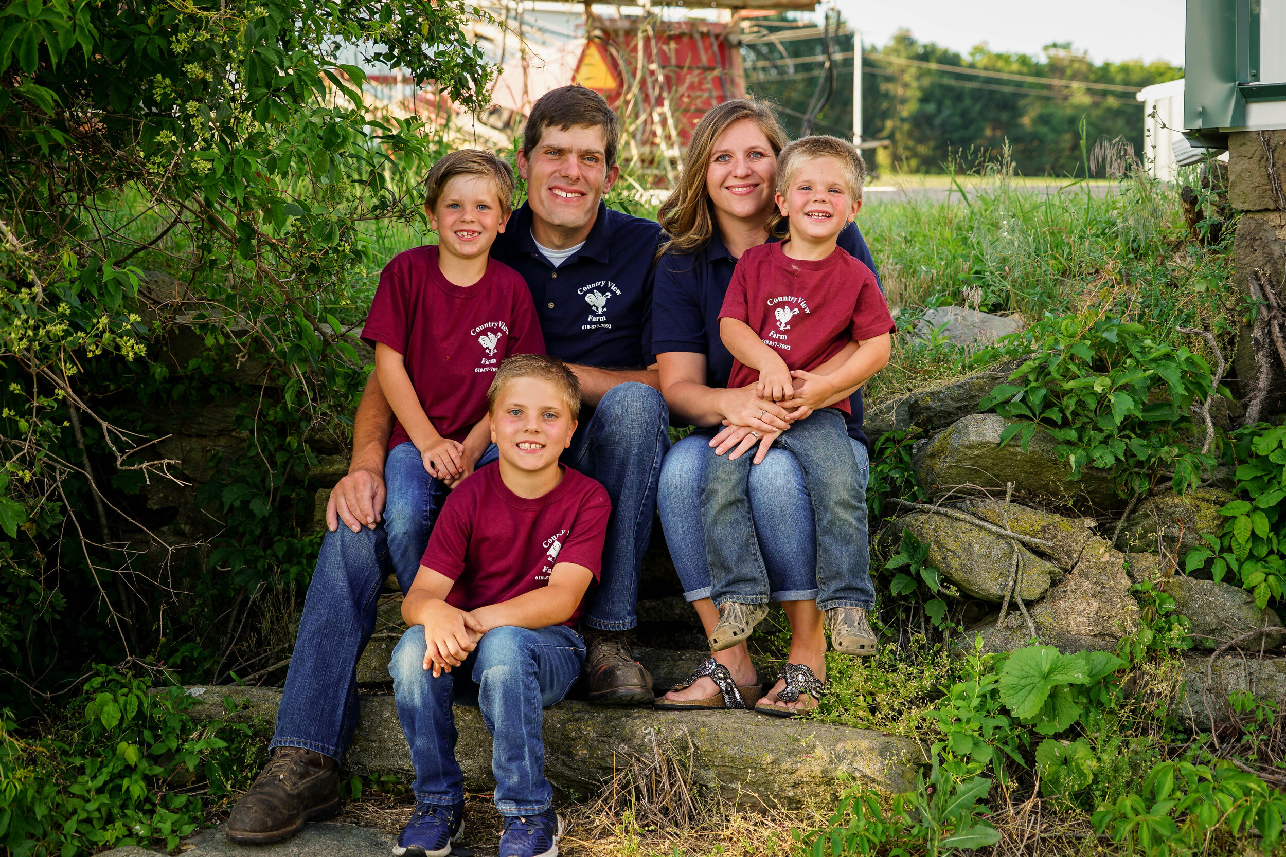 Family portrait of a smiling family sitting on stone steps surrounded by greenery, wearing matching shirts from Country View Farm, capturing a warm, outdoor moment.