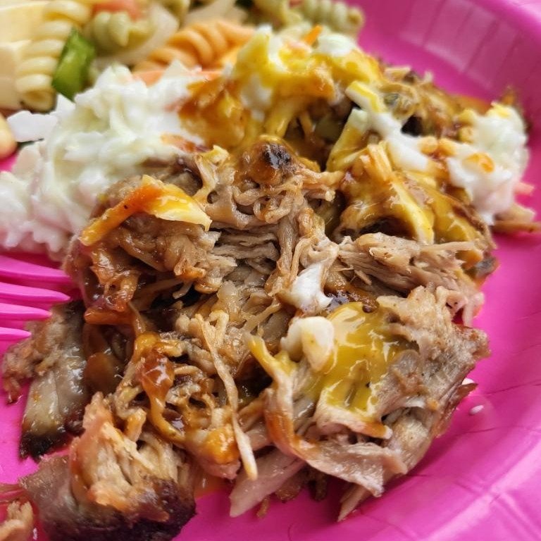 Pulled pork topped with melted cheese and barbecue sauce, served on a pink plate alongside coleslaw and pasta salad.