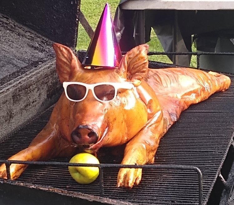 A whole roasted pig wearing sunglasses and a party hat, resting on a grill with an apple in front, symbolizing a festive barbecue or celebration.