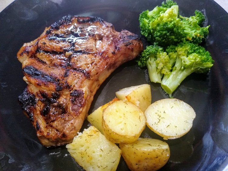 Grilled pork chop served with steamed broccoli and roasted potatoes on a black plate, showcasing a balanced meal rich in protein and vegetables.